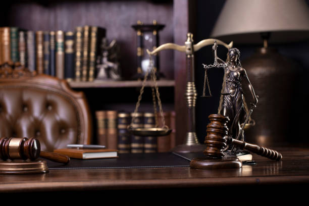 Themis statue, gavel and scale on brown desk. Legal books in the bookshelf as background.